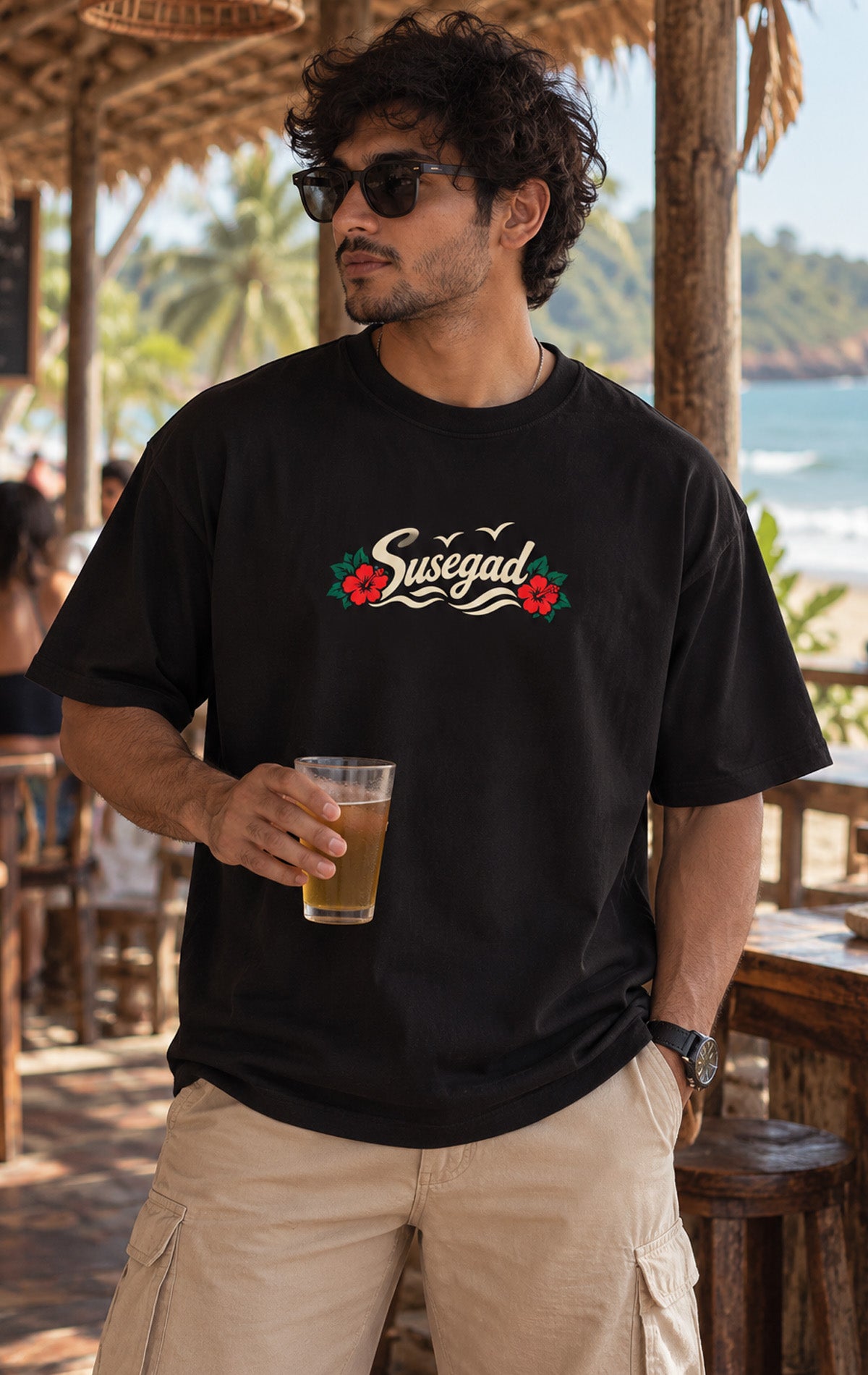 Black oversized Oi Patrao t-shirt with Susegad script and red hibiscus flowers chest print, worn at a Goa beach shack